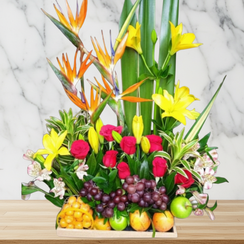 Arreglo floral con frutas tropicales sobre mesa de madera Arreglo de flores amarillas, naranjas y rojas combinado con frutas tropicales como uvas, manzanas y naranjas sobre una base de madera, con fondo de pared de mármol claro