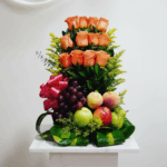 Arreglo vertical de rosas naranjas combinado con frutas frescas como uvas moradas, manzanas verdes y rojas y duraznos, decorado con hojas verdes y un lazo rosa sobre pedestal blanco