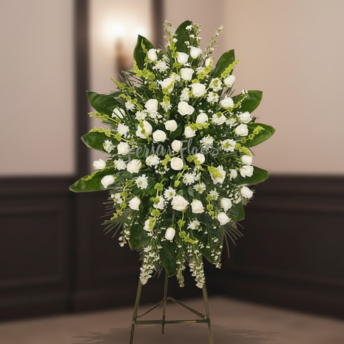 Arreglo funerario grande con rosas blancas y follaje verde sobre caballete metálico en interior