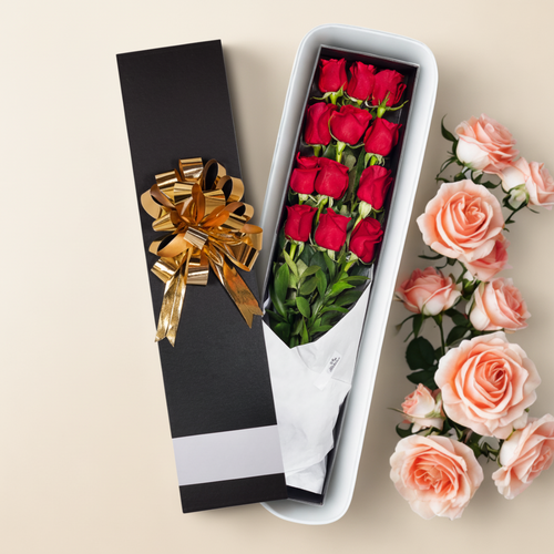 Caja de regalo alargada negra con lazo dorado que contiene un ramo de rosas rojas envueltas en papel blanco, junto a rosas rosadas sobre fondo beige