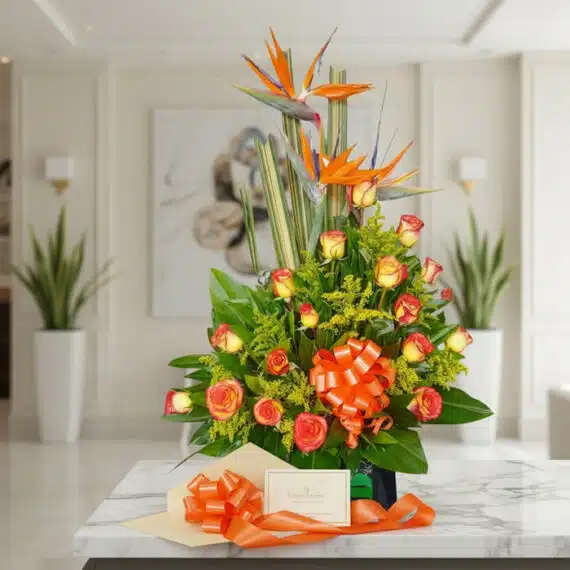 Arreglo floral exótico Esplendor con rosas naranjas y aves del paraíso, presentado en un entorno de lujo con una cinta decora
