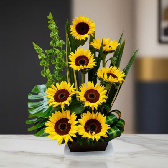 Arreglo floral premium con girasoles Bengala, hojas verdes y follaje, sobre una mesa de mármol en un lobby de hotel boutique.
