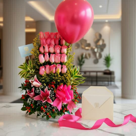 Arreglo floral elegante con rosas rosadas, lirios, follaje verde, globo fucsia, cinta de satén y sobre en mesa de mármol.