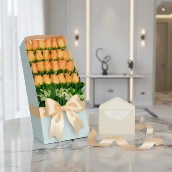 Caja de 24 rosas salmón frescas en empaque azul con lazo satinado, tarjeta de regalo y cinta sobre mármol.