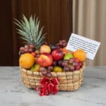 Canasta de frutas Musca con piña, uvas, manzanas, naranjas y duraznos, presentada en un entorno elegante con fondo de madera