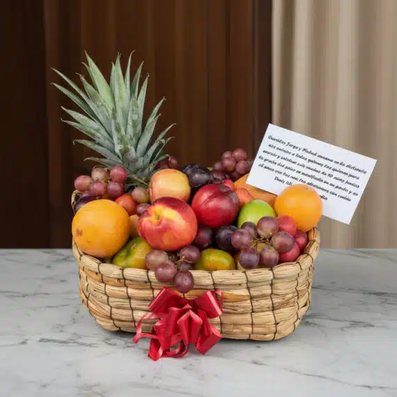 Canasta de frutas Musca con piña, uvas, manzanas, naranjas y duraznos, presentada en un entorno elegante con fondo de madera