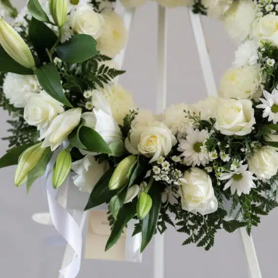 Corona fúnebre 'Homenaje Sincero' con lirios, rosas y crisantemos blancos, follaje verde, rocío sutil, cinta satinada y sobre