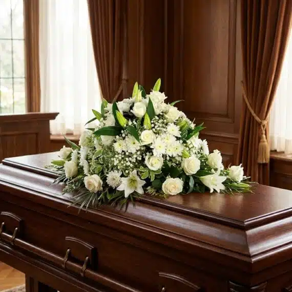 Cubre caja fúnebre solemne con rosas blancas, lirios y follaje verde sobre un ataúd de madera oscura en un entorno elegante y