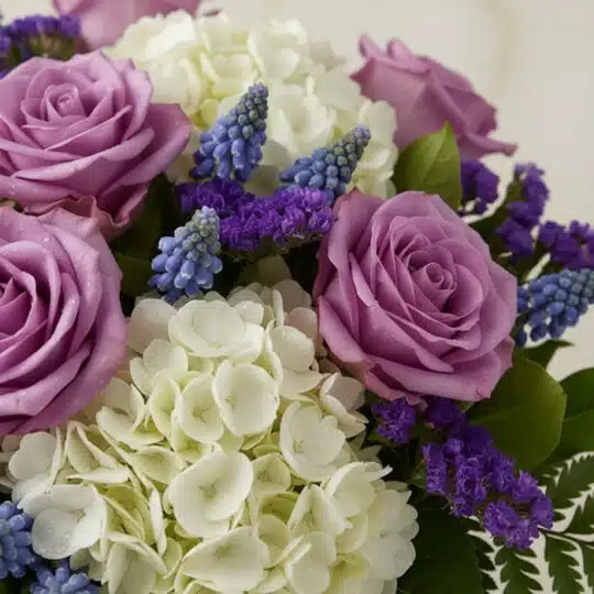 Primer plano extremo de Jarron Floral VITTORIA con rosas lilas, hortensias blancas, muscari azul y statice morado, mostrando