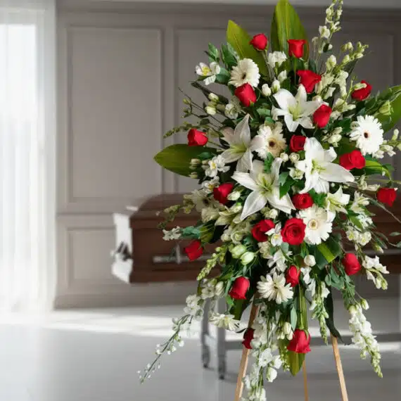 Pedestal fúnebre Adoración con lirios blancos, rosas rojas y gerberas blancas en sala de velación moderna, transmitiendo resp